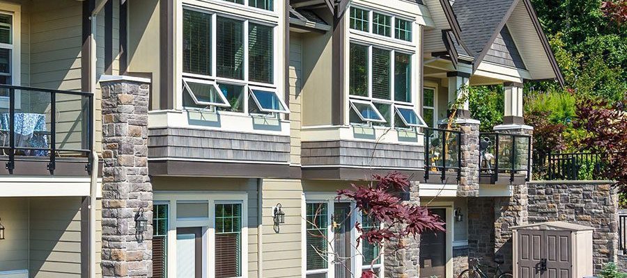 A modern townhouse with beige siding, stone accents, large windows, and a decorative balcony overlooks a paved patio area with outdoor furniture and plants.