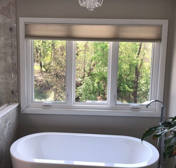 A bathroom with a white freestanding bathtub, a plant to the side, large window overlooking trees, and a glass-enclosed shower on the left.