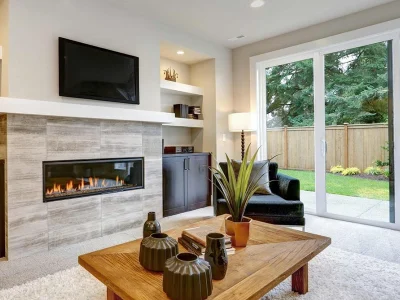 A modern living room with a wall-mounted TV above a fireplace, a sliding glass door opening to a garden, a wooden coffee table with decorative items, and a black armchair beside a plant.