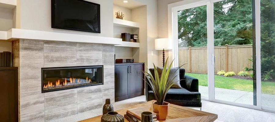 A modern living room with a wall-mounted TV above a fireplace, a sliding glass door opening to a garden, a wooden coffee table with decorative items, and a black armchair beside a plant.