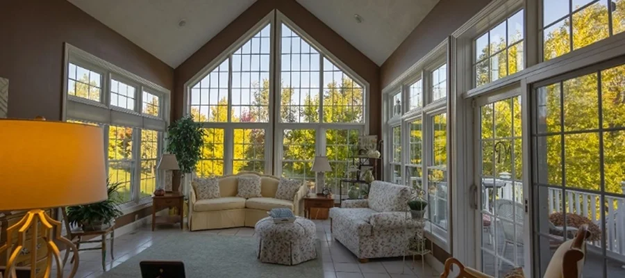A sunlit room with large floor-to-ceiling windows, furnished with beige couches, armchairs, a lamp, and indoor plants, featuring a view of a green, wooded area outside.