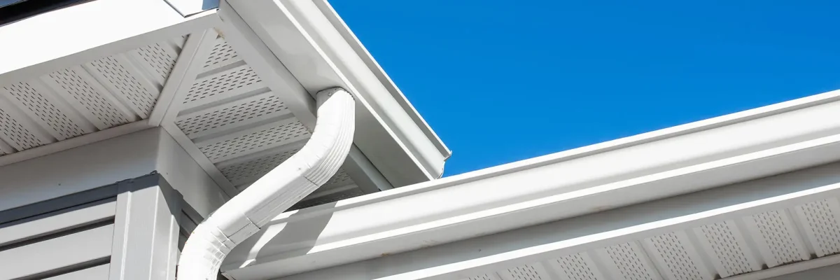Close-up view of a white rain gutter and downspout on the corner of a building's roof against a clear blue sky.