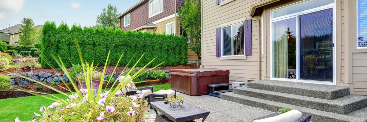 A backyard with patio furniture, a hot tub, potted plants, and steps leading to a sliding glass door of a two-story house.